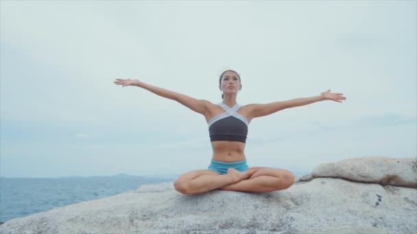 Mujer levantando las manos por encima de la cabeza y meditando mientras está en roca cerca del océano — Vídeos de Stock