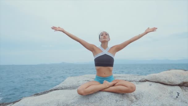 Mujer levantando las manos por encima de la cabeza y meditando mientras está en roca cerca del océano — Vídeos de Stock