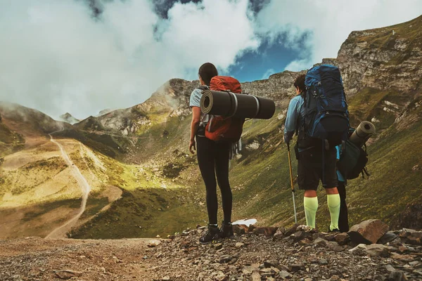 Hiking team with heavy backpacks stands on top of a mountain and enjoying camping valley view