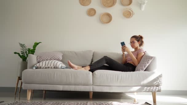 Jeune femme détendue utilisant un téléphone intelligent surfer sur les médias sociaux, vérifier les nouvelles, jouer à des jeux mobiles ou envoyer des messages texte assis sur le canapé — Video