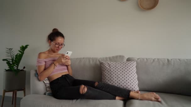 Jovem mulher celebra sua vitória, olhando telefone inteligente no sofá — Vídeo de Stock