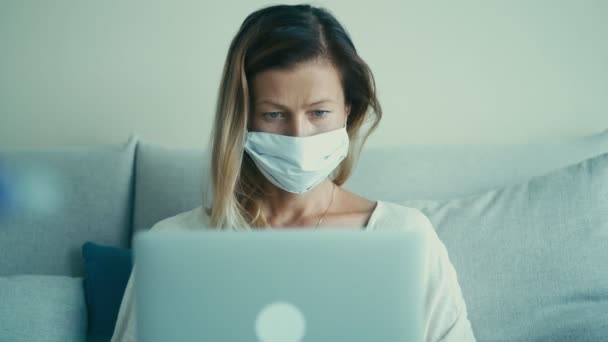 Kobieta biznesu w jednorazowej masce medycznej jest wpisując na klawiaturze laptopa na kanapie — Wideo stockowe