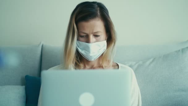 Kobieta biznesu w jednorazowej masce medycznej jest wpisując na klawiaturze laptopa na kanapie — Wideo stockowe
