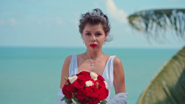 Krásná nevěsta s kyticí růží stojí a dívá se do kamery na pozadí moře v její svatební den — Stock video