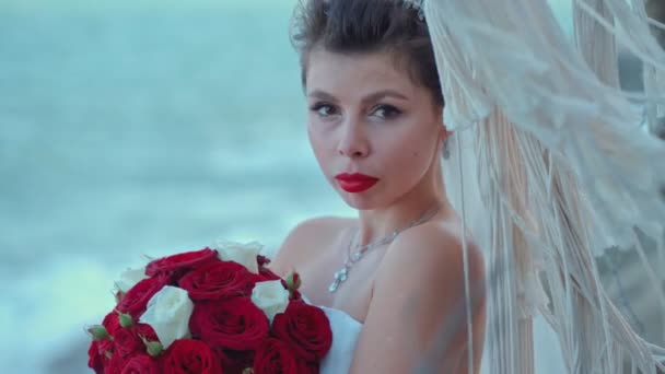 Una hermosa novia con un ramo de rosas está mirando a la cámara contra el fondo del mar en el día de su boda — Vídeos de Stock