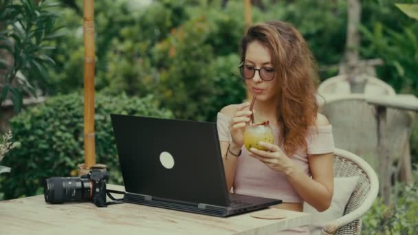Молода жінка-фотограф п'є коктейль під час роботи зі своїм ноутбуком у відкритому кафе — стокове відео