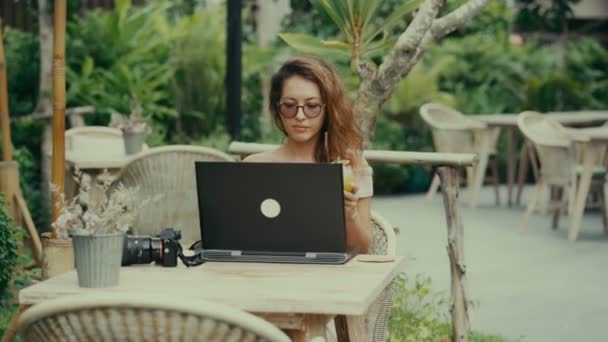 A young woman photographer drinks a cocktail while working with her laptop in an outdoor cafe — Stock Video