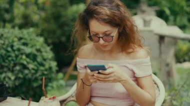 Genç bir kadın güneşli bir günde açık kahve dükkanında akıllı telefondan mesaj atıyor.