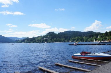 Almanya 'da Titisee Gölü ve Kara Orman 'ın ormanlık Dağları manzarası, göldeki tekneler, mavi gökyüzü ve beyaz bulutlarla güneşli gün
