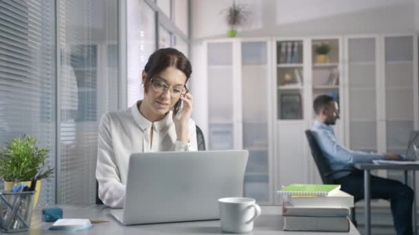 Wanita cantik kasual di meja menggunakan laptop menjawab panggilan berbicara di telepon — Stok Video