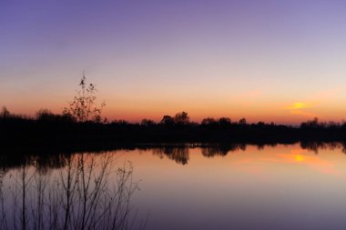 Mavi zaman akşam gökyüzü manzarası, önünde ağaçlar ve göl var. Mavi zaman gün batımı. Mükemmel deniz mavisi ve turuncu renkler. Düşük ışıkta çekilen fotoğraf.