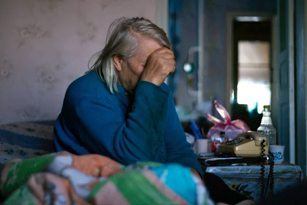 Yaşlı, gri saçlı kadın elini yüzünü kapadı. Kadın üzgün. Köyde fakir bir hayat. Yaşlılık iyi değil. Düşük ışık fotoğrafı. 