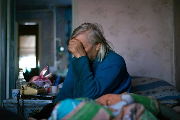 Yaşlı, gri saçlı kadın elini yüzünü kapadı. Kadın üzgün. Köyde fakir bir hayat. Yaşlılık iyi değil. Düşük ışık fotoğrafı. 