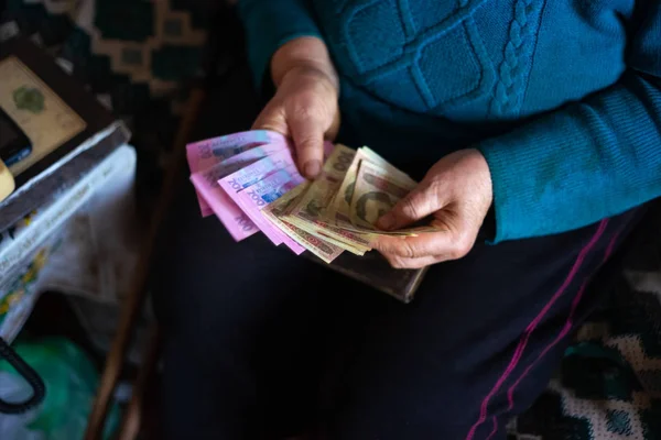 Yaşlı, gri saçlı kadın Ukrayna 'nın Hryvna parasını elinde tutuyor. Kadın üzgün. Köyde fakir bir hayat. Yaşlılık iyi değil. Düşük ışık fotoğrafı.