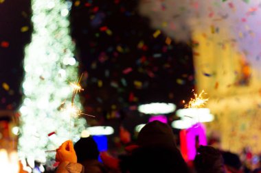 Ana meydanda yeni yıl kutlaması. Bir sürü duman, sis, ışıklar, selam ve konfeti. Bulanık arkaplan. Ön planda eldivenle Bengali ateşi. Çerçevenin sağ tarafında mavi ışık olan konser sahnesi.