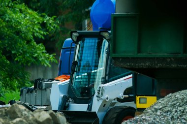 Öndeki moloz ve toprak yığını. Arka planda küçük bir buldozer yükleyicisi ve bir kamyon var.