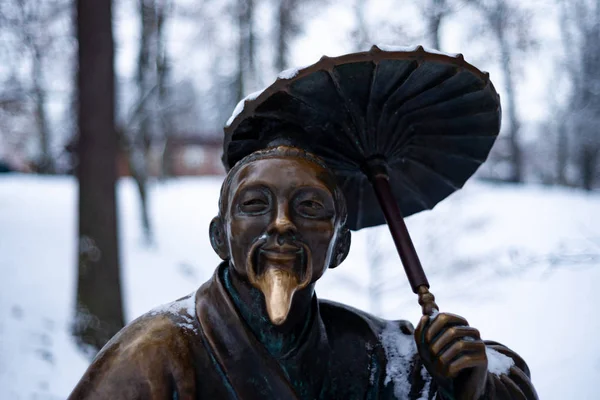 Bronze monument of a chinese old man with umbrella in winter snow park. Closeup with blurred background.