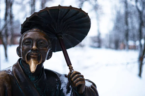 Bronze monument of a chinese old man with umbrella in winter snow park. Closeup with blurred background.