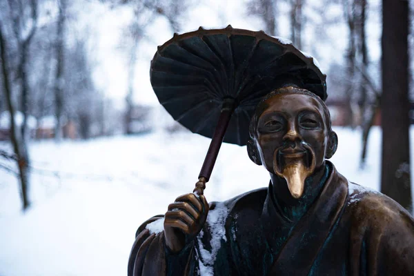 Bronze monument of a chinese old man with umbrella in winter snow park. Closeup with blurred background.