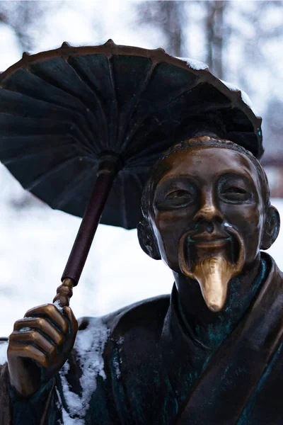 Bronze monument of a chinese old man with umbrella in winter snow park. Closeup with blurred background.