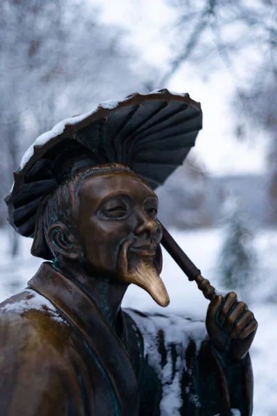 Bronze monument of a chinese old man with umbrella in winter snow park. Closeup with blurred background.