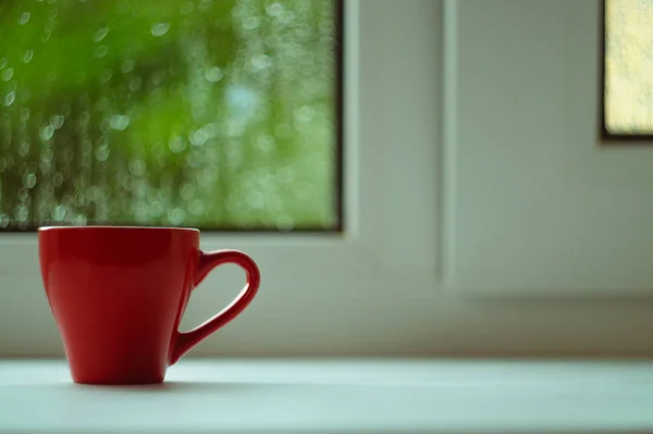 The red espresso cup on white windowsill. On background window and rain. Cup located at left of frame.