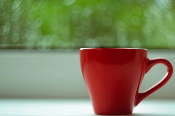 The red espresso cup on white windowsill. On background window and rain. Cup located at right of frame.