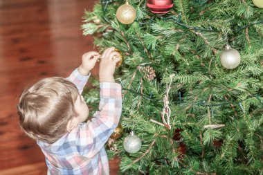 Sevimli küçük bir çocuk oturma odasında büyük bir Noel ağacına dekoratif bir top yerleştirir
