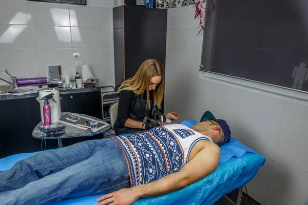 Blonde tattooist girl works in her small studio, tattooing the a