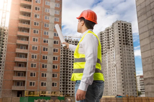 Architetto maschio in piedi sul cantiere e che punta su edifici in costruzione con cianografie — Foto Stock
