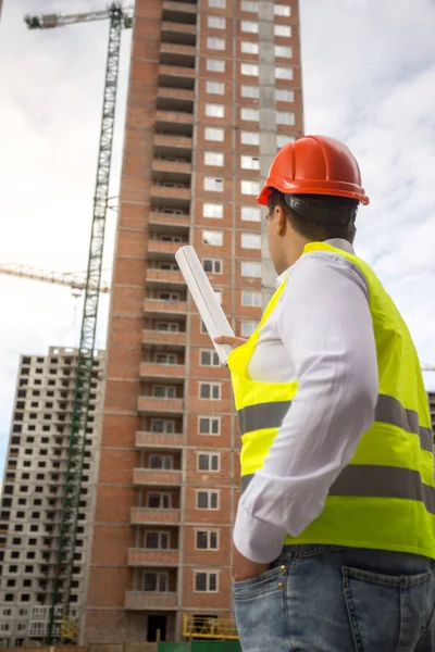 Rear view image of male architect pointing at buildings under construction with roll of blueprints — Stock Photo, Image