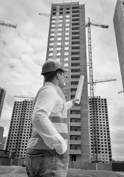 Black and white image of male architect standing on construction site and pointing at building under construction