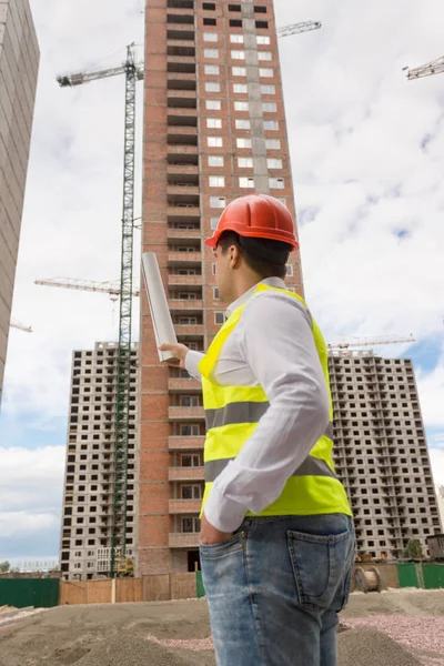 Rear view image of male architect in hardhat holding blueprints and pointing at construction site — Stock Photo, Image