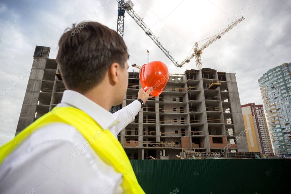 Rear view image of male achitect pointing with red hardhat on new building under construction