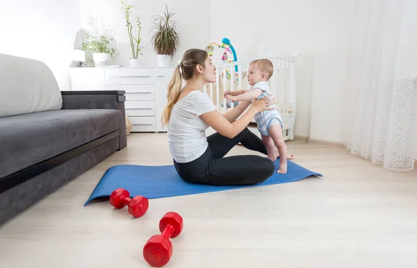 Schöne junge Frau macht Fitnessübungen mit ihrem kleinen Jungen auf dem Boden zu Hause — Stockfoto