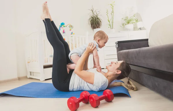 Hermosa mujer joven sosteniendo a su bebé niño y haciendo ejercicio en el suelo en casa — Foto de Stock