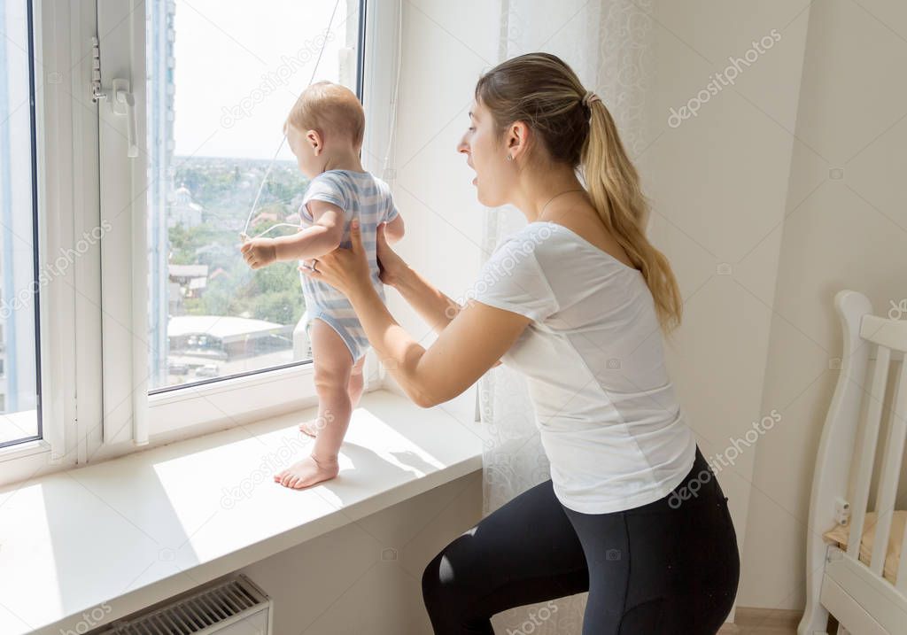 Young mother catching and holding her baby son standing on sindowsil and looking out of the window