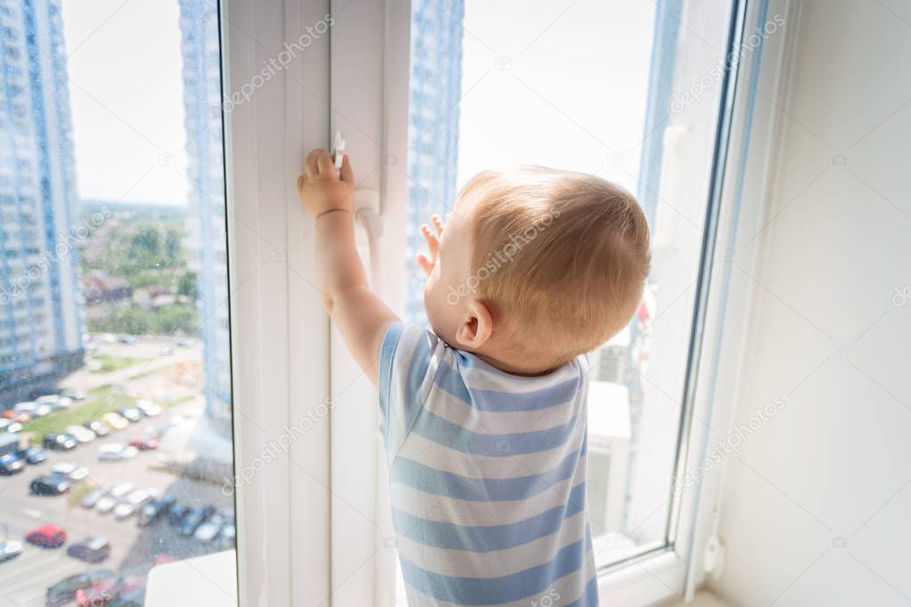 9 months old baby boy standing on windowsill and trying to open the window. Bbay in danger