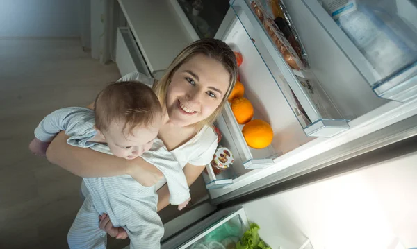 Portrét mladé matky drží její chlapeček a stojící na otevřené lednice na kuchyň v noci — Stock fotografie