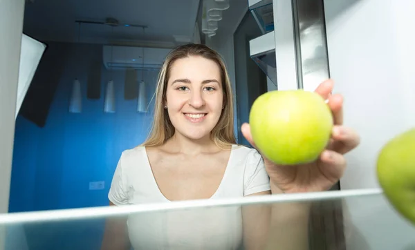 Retrato de uma jovem sorrindo de pijama tirando maçã verde fresca da geladeira à noite — Fotografia de Stock