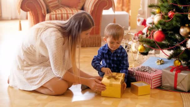 4k video of adorable toddler boy sitting on floor with mother under Christmas tree and opening gift box with presents — Stock Video