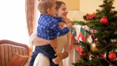 Genç Anne onu yürümeye başlayan çocuk tutan küçük oğlu ve süsleyen Noel ağacı oturma odası 4 k görüntüleri