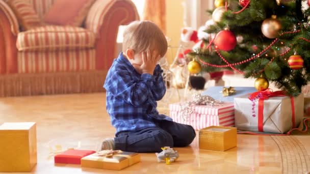 4k metraje de niño molesto sentado bajo el árbol de Navidad y llorando a causa de regalo no deseado de Santa — Vídeos de Stock