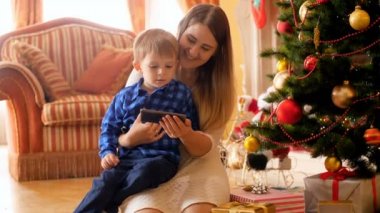 4 k video gülümseyen Genç Anne ile küçük oğlu Noel ağacının altında cep telefonunda çizgi film izlerken
