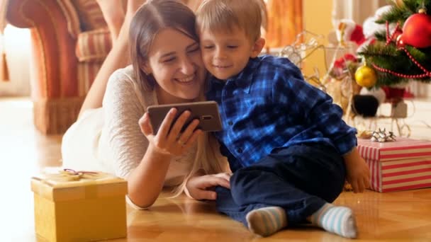 4 k wideo szczęśliwą rodzinę, leżąc na podłodze pod choinkę i oglądania inteligentny telefon — Wideo stockowe