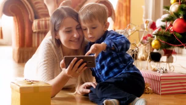 Schattig 3 jaar oude peuter jongen kijken cartoons op mobiele telefoon met jonge moeder op kerstochtend — Stockvideo