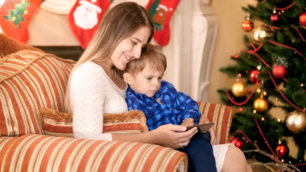 Feliz madre sonriente sentada en el sillón con su hijo pequeño y viendo video en el teléfono inteligente. Árbol de Navidad y chimenea decorada para vacaciones de invierno en el fondo — Vídeos de Stock