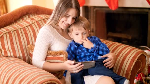 3-летний симпатичный мальчик сидит с мамой в кресле в гостиной и смотрит видео на экране мобильного телефона — стоковое видео