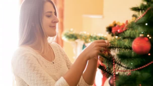 カラフルなつまらないものとビーズ付きのリビング ルームでクリスマス ツリーを飾る美しい笑顔の女性の肖像画 — ストック動画