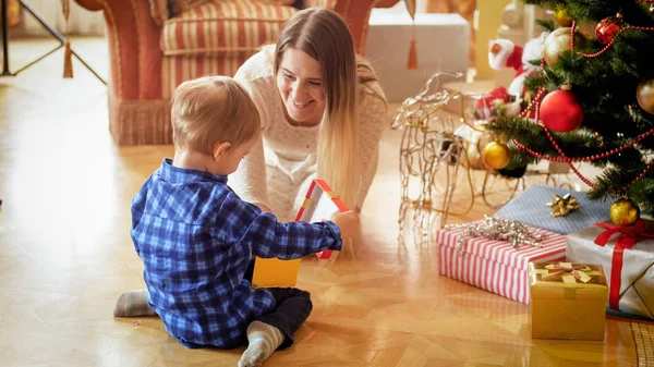 Мальчик сидит на полу с матерью и открывает коробку с рождественским подарком — стоковое фото